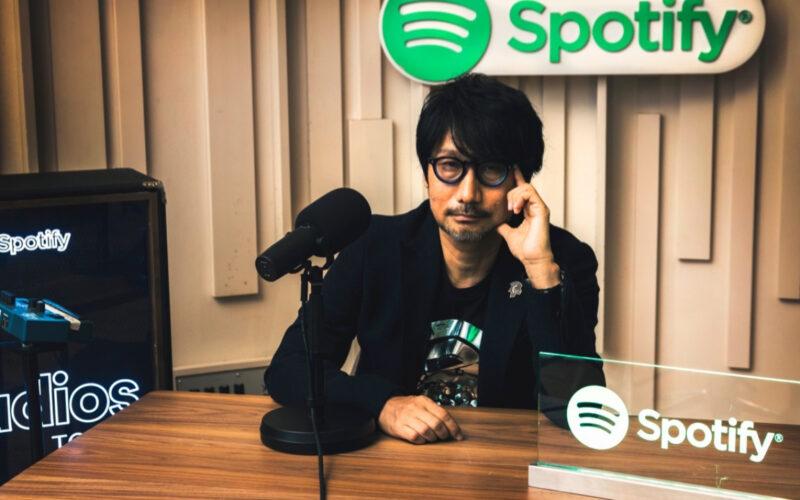 Hideo Kojima yanında ve altında Spotify logolarıyla oturuyor