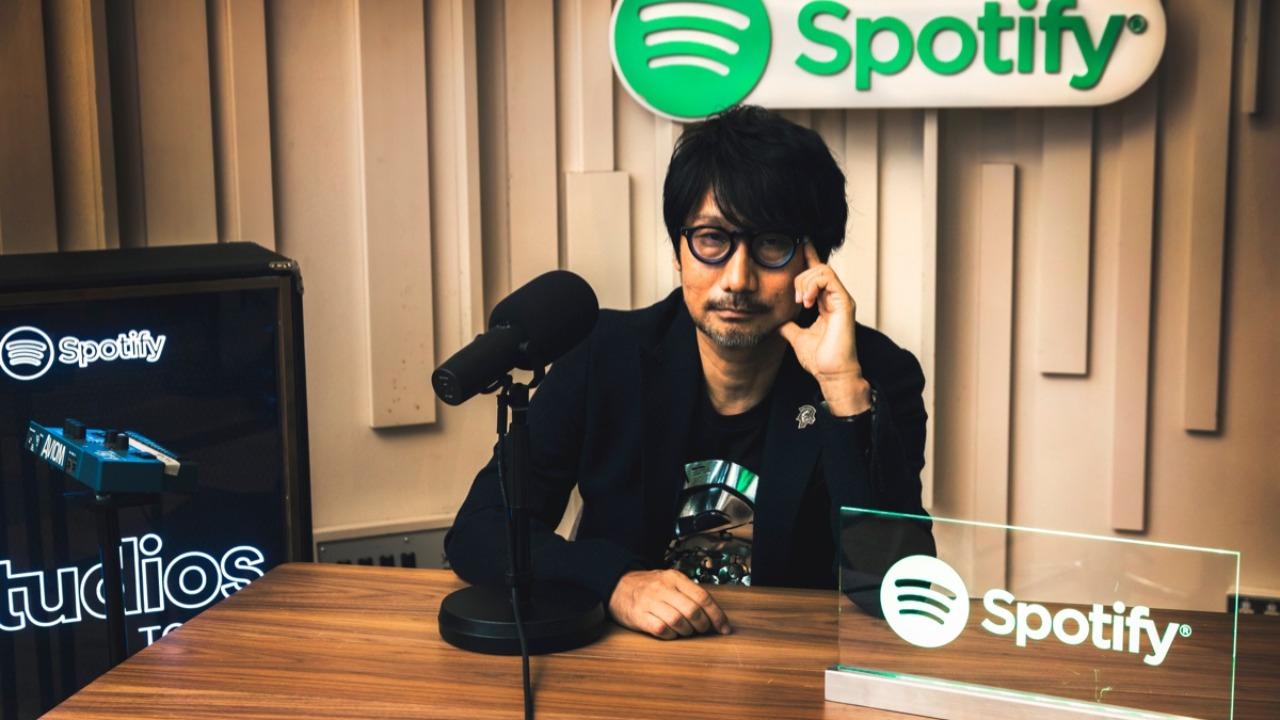 Hideo Kojima yanında ve altında Spotify logolarıyla oturuyor