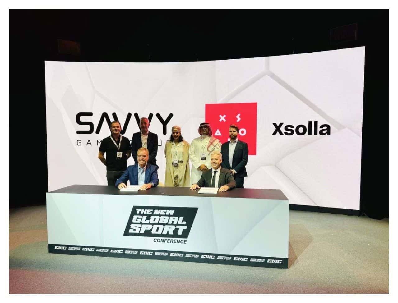 Xsolla CFO Chris Hewish and Savvy Group CEO Brian Ward on a stage signing MoU with five other person standing behind them presumably from the Saudi Arabia gaming industry